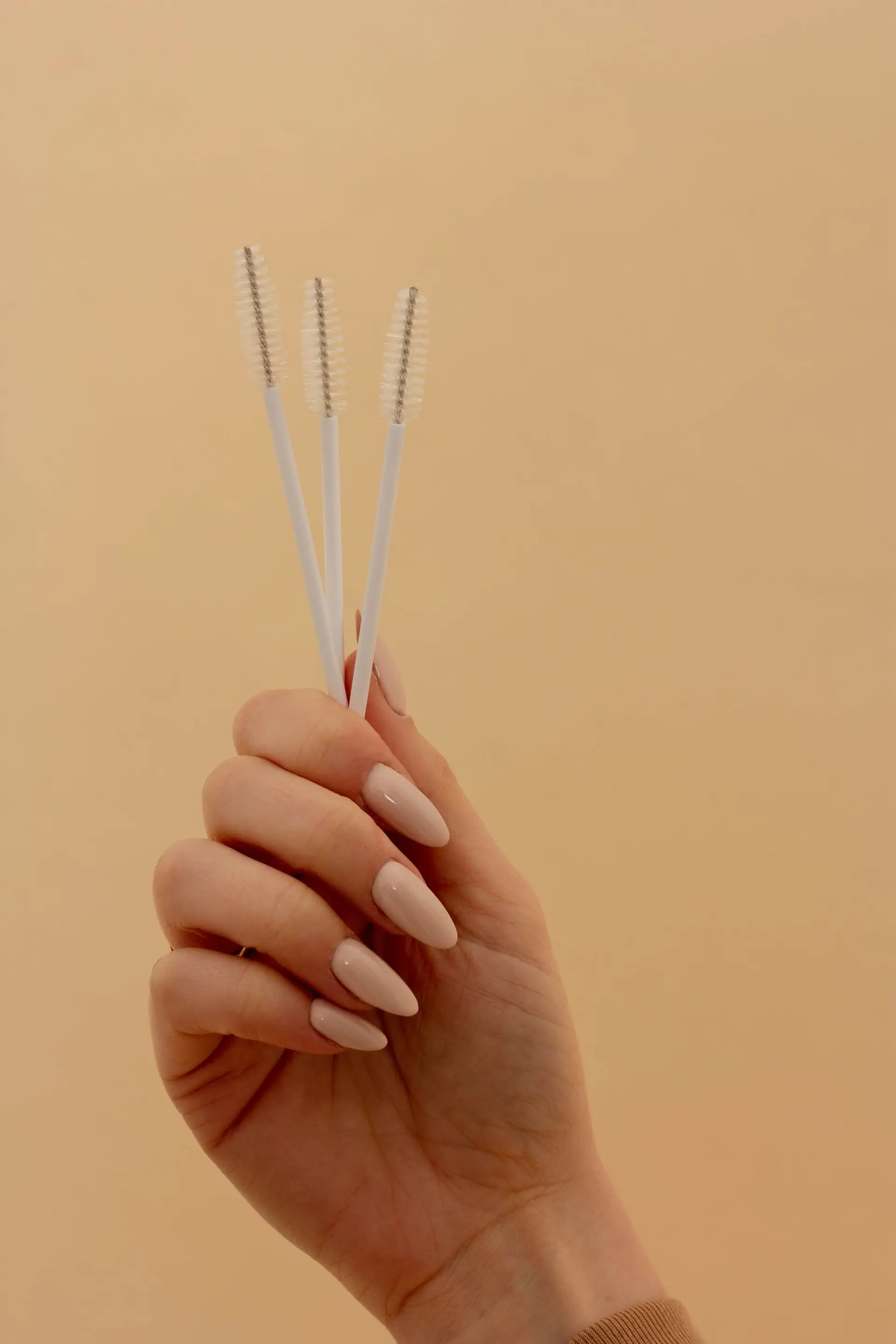 A hand holding three brow spoolies against a soft peach background